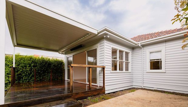 Rear exterior view of a single-storey weatherboard house with a covered timber deck, pergola-style roof extension, timber balustrade, white-painted cladding, and traditional multi-pane windows. The scene appears to be a compact courtyard or backyard entertaining area.