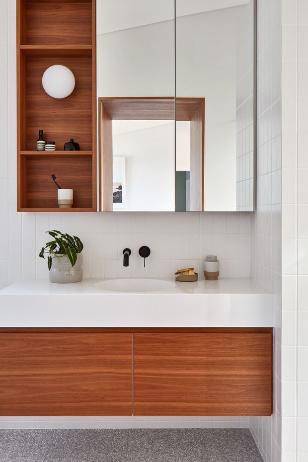 A bright, contemporary bathroom vanity featuring a white integrated basin and benchtop, warm timber wall shelving and floating cabinetry, large mirrored cabinet doors, white square wall tiles, and matte black tapware. Minimal styling includes a potted plant and small bathroom accessories. A doorway and adjoining interior are reflected in the mirror.
