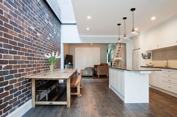 Open-plan kitchen, dining and living area in a renovated brick-lined interior. A white island kitchen with shaker-style cabinetry, tiled splashback and pendant lighting sits beside a timber dining table, while a high exposed brick wall and dark timber flooring add character. The living area and staircase are visible beyond.