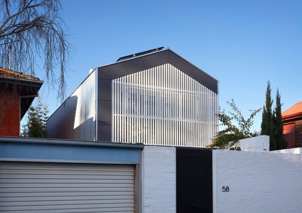 Street-facing view of a contemporary residential building behind a white masonry boundary wall. The building has a steep gabled form clad in dark metal panels, with a prominent vertical white slatted screen across the front facade. A garage door and pedestrian gate are visible in the foreground, with neighbouring homes and landscaping at the edges.
