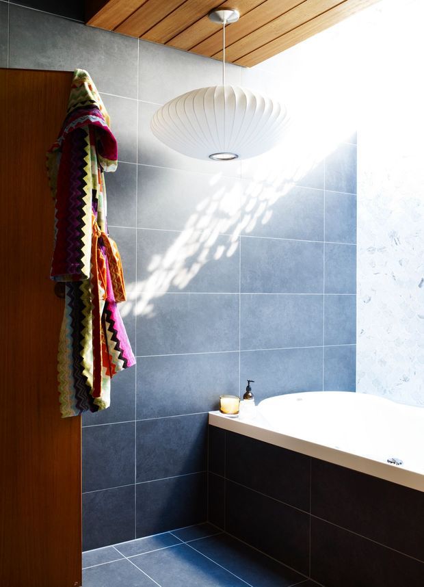A contemporary bathroom with large blue-grey tiled walls and floor, a white built-in bathtub with dark tiled surround, a timber slatted ceiling, and a sculptural white pendant light. A timber door and colourful patterned towels add warmth and contrast, while strong daylight creates dappled shadows across the tiles.