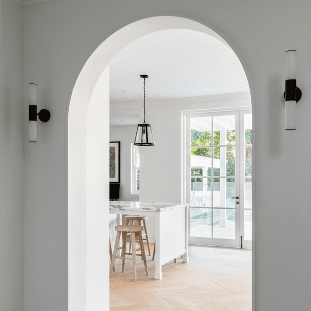 Bright, refined interior view through a large arched opening into a white kitchen. The kitchen features a pale stone island with timber stools, a black pendant light, white cabinetry, herringbone timber flooring, and full-height glazed French doors opening to greenery outside. Slim black-and-glass wall sconces flank the archway.