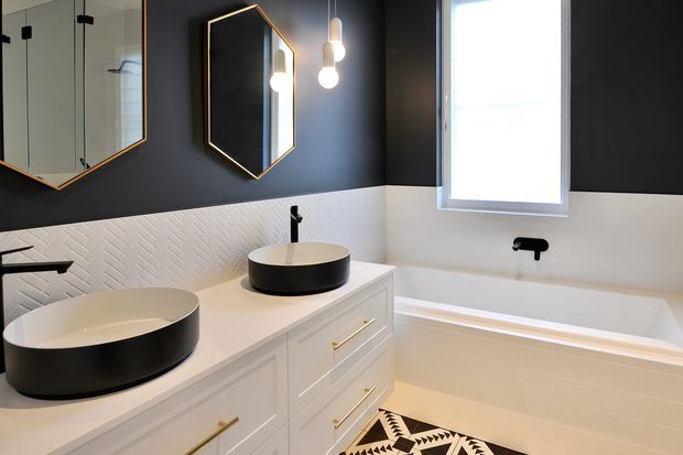 A contemporary bathroom featuring a double white vanity with black vessel basins, white cabinetry with brass pulls, a white textured herringbone tile backsplash, and a freestanding-style built-in bathtub. The room has dark charcoal walls, geometric brass-framed mirrors, black tapware, warm pendant lighting, and a patterned floor mat.
