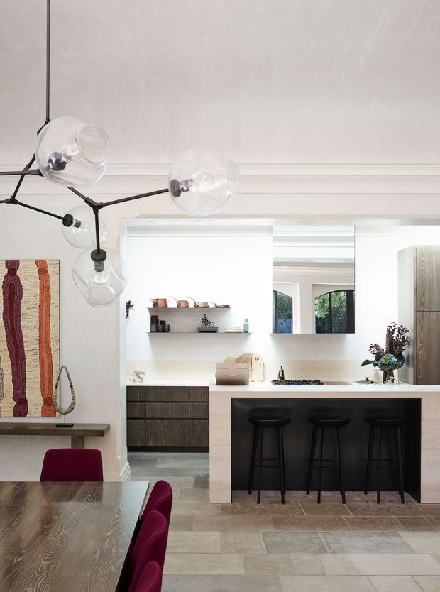 A bright, refined open-plan kitchen and dining area with a pale neutral palette. The kitchen features a light stone island with a black front panel, dark timber cabinetry, open shelving, a large window, and black bar stools. In the foreground is a timber dining table with burgundy upholstered chairs, while a sculptural glass-bulb pendant light hangs overhead. Light stone or concrete-look floor tiles, wall moulding, and a framed textile artwork complete the space.