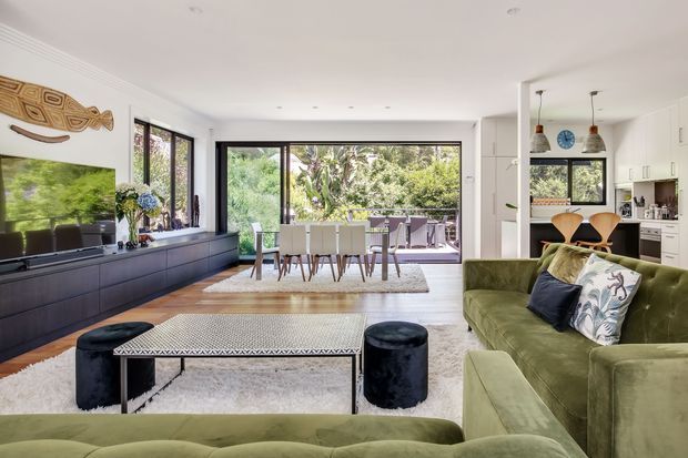 Open-plan contemporary living, dining and kitchen space with a layered green and neutral palette. The room features a large green sectional sofa, black upholstered ottomans, a patterned rectangular coffee table, a wall-mounted television above dark built-in cabinetry, and a dining table positioned beside expansive windows and glazed doors opening to a leafy outdoor area. A compact white kitchen with an island and timber stools is visible at the rear.