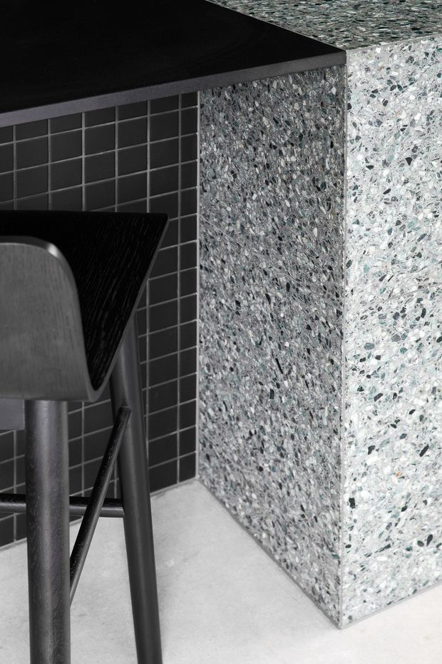 Close-up detail of a dark, minimalist counter or island corner with a black benchtop, terrazzo-clad side panels, a charcoal square-tile wall, and a partially visible black and grey bar stool. The image emphasizes material textures and crisp geometric junctions.