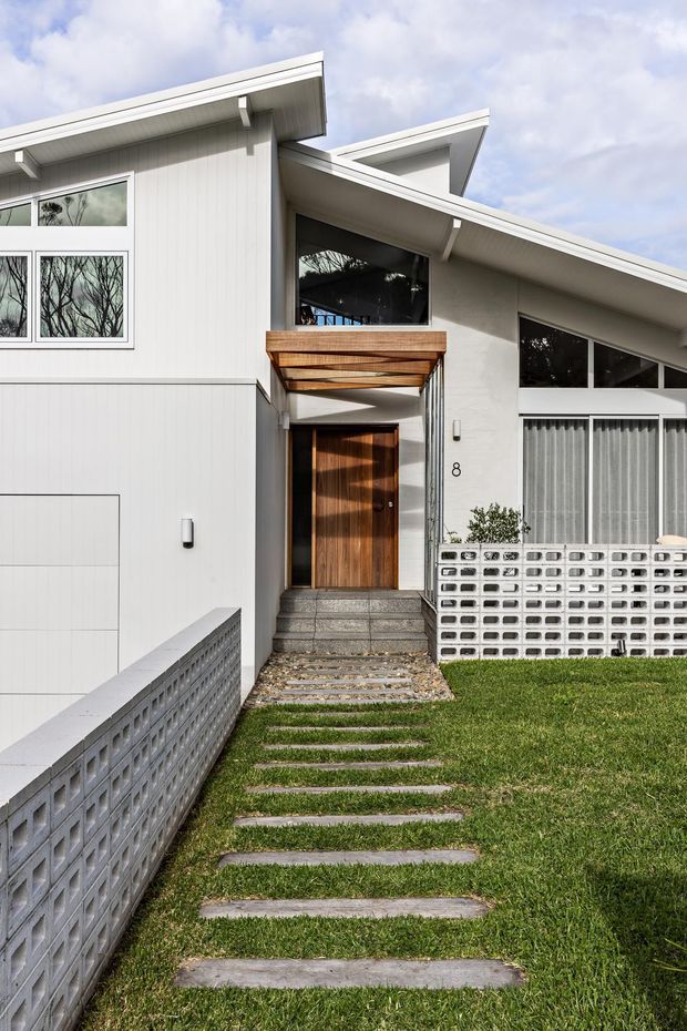 Exterior view of a contemporary residential entrance approached by a stepping-stone path through lawn. The house has overlapping asymmetrical skillion roofs, white vertical cladding and rendered walls, large glazed openings, timber entry door and canopy, and a distinctive white breeze-block boundary wall.
