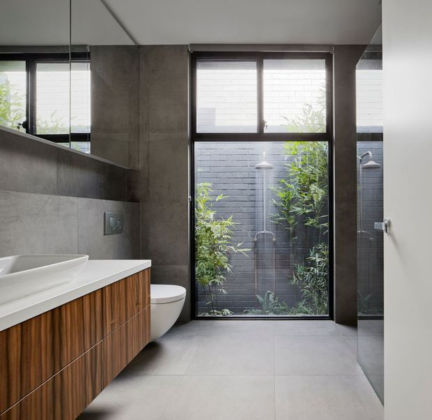 Contemporary bathroom with a wall-mounted timber vanity, white integrated basin, concealed toilet, large grey tiled surfaces, dark-framed windows and a glass shower enclosure. The bathroom opens toward a planted private courtyard, bringing greenery and daylight into the space. Clean-lined fixtures and restrained neutral finishes create a calm, refined atmosphere.
