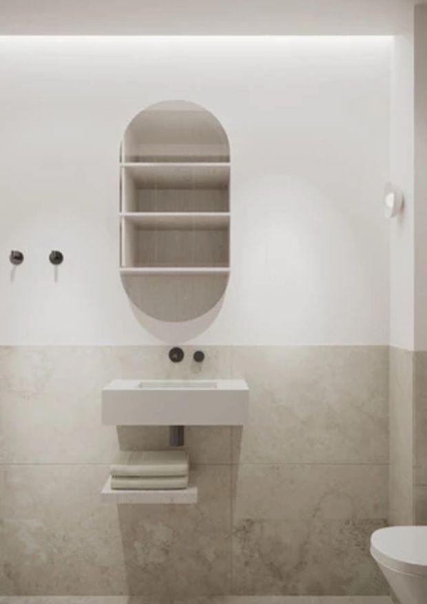 A compact, minimalist bathroom with pale marble-look wall tiles, a wall-mounted white basin, exposed plumbing, and a partially visible toilet. Above the basin is a distinctive rounded-rectangle recessed shelving niche with light timber-toned backing. The room has white painted upper walls, understated dark wall fittings, and concealed linear lighting near the ceiling.