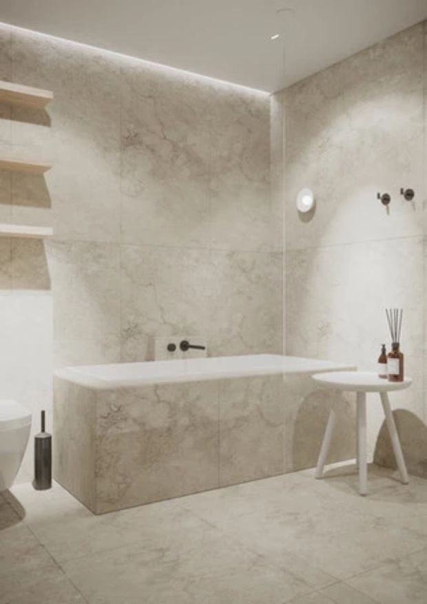A refined, minimalist bathroom clad in pale marble-look stone across the walls, bathtub surround, and floor. The room includes a sculptural freestanding bathtub, wall-mounted bath fittings, integrated open shelving, a small white side table with fragrance bottles, and concealed warm LED lighting along the ceiling perimeter.