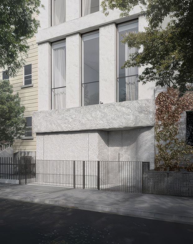 Street-facing view of a contemporary multi-storey residential facade. The building is finished in pale textured render or concrete-like surfaces, with tall recessed windows, slim dark frames, a projecting upper-level balcony or solid ledge, and a dark metal pedestrian and vehicle gate. Mature street trees partially screen the facade, with a neighbouring weatherboard building visible at left.