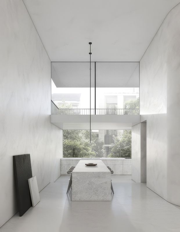 A minimalist double-height dining room with white sculptural walls, a long pale stone dining table, dark slim chairs, and a linear pendant suspended through the tall void. Full-height glazing at the rear overlooks an internal balcony and leafy urban surroundings, creating a bright, monochromatic and gallery-like space.