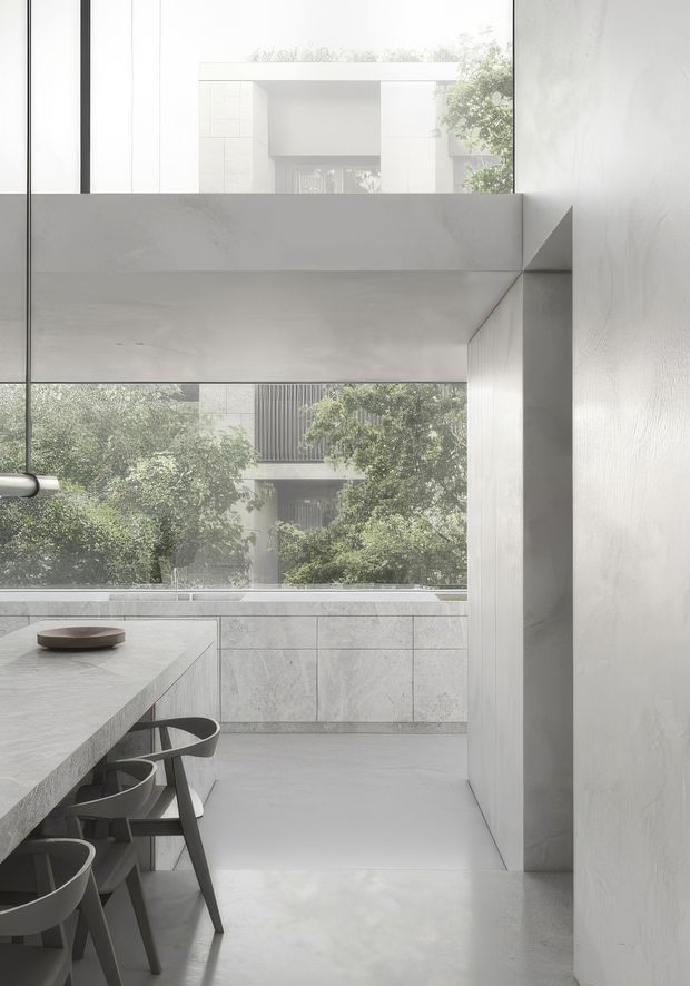 A minimalist, light-filled kitchen with a long stone island and dark sculptural stools. Pale grey stone cabinetry and surfaces extend across the rear wall, while a large glazed opening frames leafy vegetation and a neighbouring contemporary building. A broad concrete or plaster beam and crisp wall junctions create a strong geometric architectural composition.