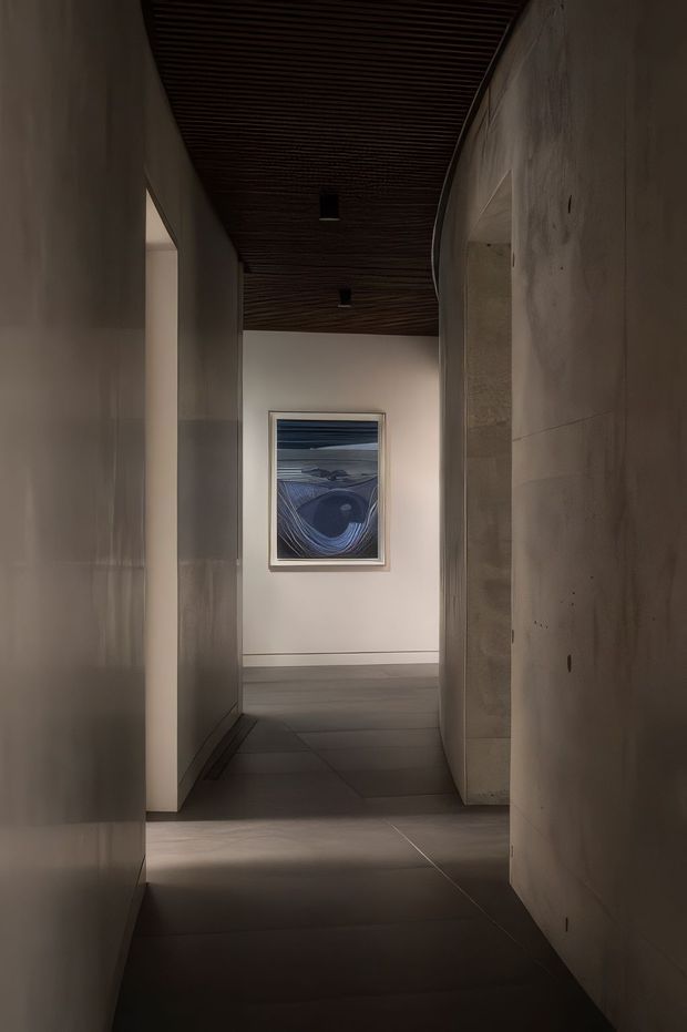 A dimly lit, narrow interior hallway with smooth pale stone or plaster walls, large dark grey floor tiles, and a curved wall opening on the right. The ceiling features dark timber slatted lining with recessed black downlights. An abstract blue-toned artwork is mounted at the far end, creating a restrained, gallery-like atmosphere.