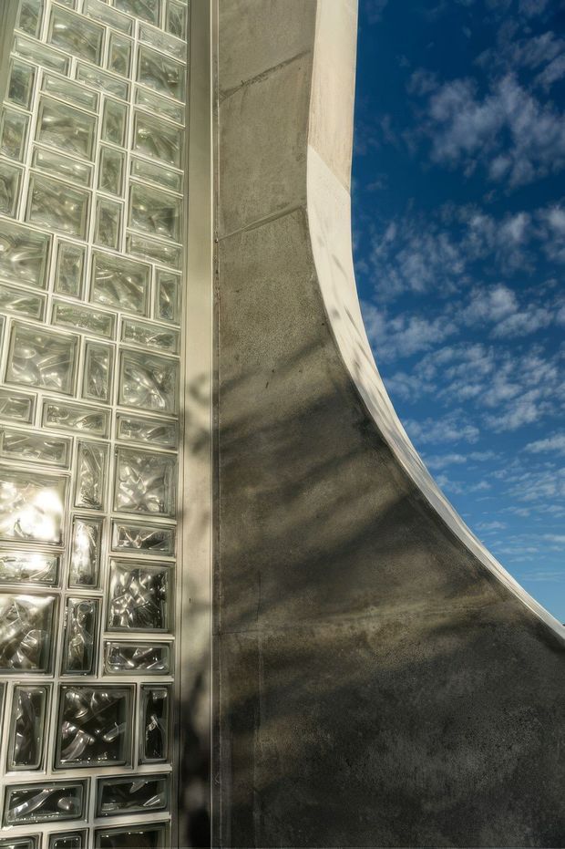 Close-up upward-looking architectural exterior detail showing a tall wall of translucent glass blocks beside a sweeping curved dark concrete facade or roof edge. Strong sunlight creates shadows across the textured surface, with blue sky and scattered clouds visible to the right.
