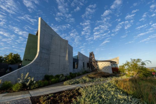 Exterior architectural photograph of a sculptural modern concrete residence, featuring sweeping curved and angular wall forms, a projecting flat roof, dark recessed glazing, and landscaped xeriscape planting in the foreground. The building sits in an open suburban or semi-arid setting beneath a bright blue sky.