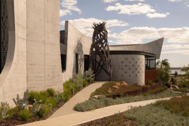 Exterior view of a sculptural contemporary concrete building set within drought-tolerant landscaped gardens. The composition features curved and angular concrete walls, narrow dark glazing, a cylindrical perforated concrete volume, and a prominent branching timber or metal sculptural screen at the entrance. Pale concrete paths weave through low grasses and groundcover, with distant landscape and sky beyond.