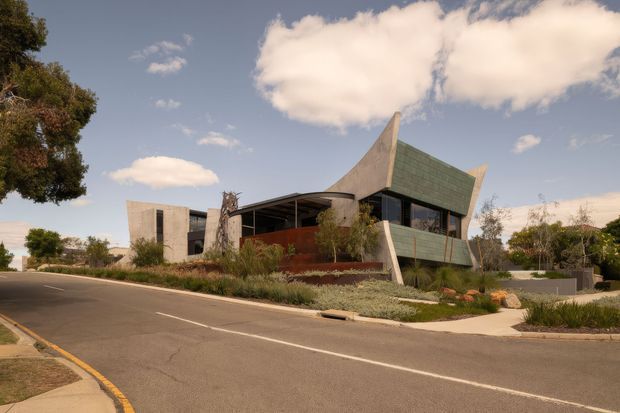 Street-level exterior view of a distinctive contemporary building with angular sculptural forms, greenish cladding, exposed concrete elements, extensive glazing, and landscaped planting around the frontage. The building sits on a suburban street beneath a bright partly cloudy sky.