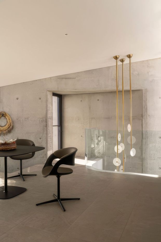 A minimalist contemporary dining area with a round black table and sculptural black chairs, set against exposed raw concrete walls and a broad concrete floor. A tall cluster of slender brass pendant lights with circular white discs hangs beside a glass balustrade overlooking a lower level. The room has high ceilings, restrained detailing, and warm natural light.