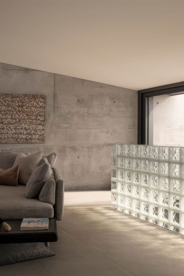 A refined contemporary living room with a low grey sectional sofa, dark coffee table, and neutral cushions. The room features exposed-looking smooth concrete wall panels, large-format concrete-toned flooring, a black-framed window, and a curved glass-block partition that diffuses strong natural light. A textured abstract artwork adds a subtle focal point.