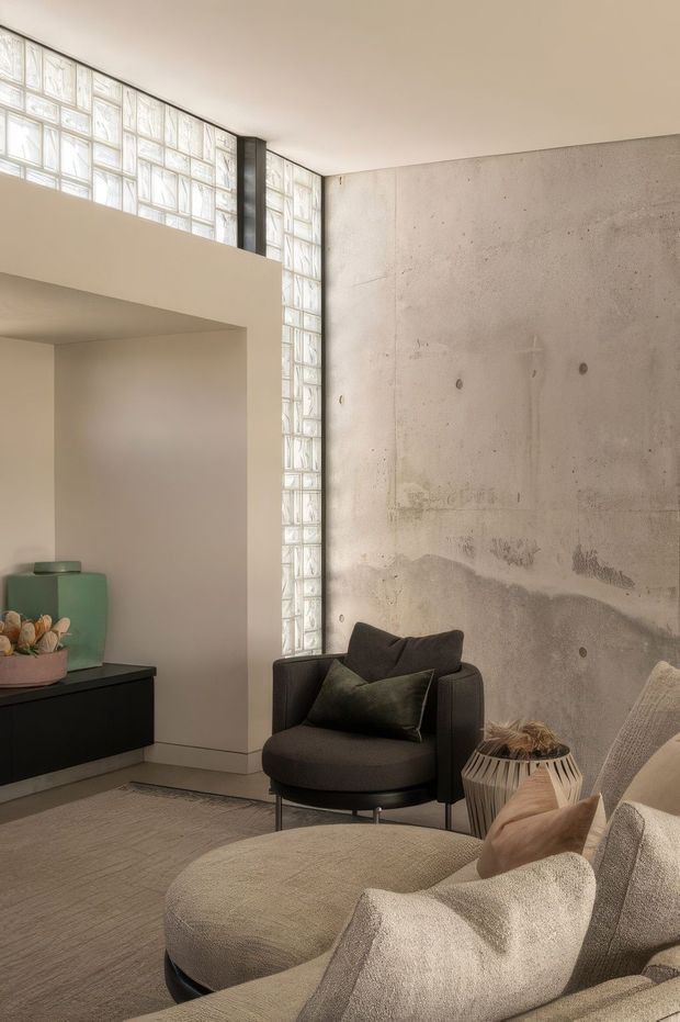 A refined contemporary living room with a curved cream sectional sofa and dark upholstered lounge chair arranged around a low ottoman. The room features a raw exposed concrete feature wall, a tall translucent glass-block window, recessed architectural niches, dark built-in cabinetry, and warm neutral finishes. Soft, indirect daylight creates a calm, understated atmosphere.