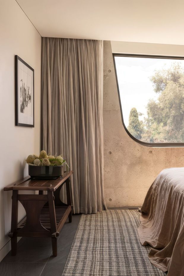 A warm, moody bedroom corner with a partially visible bed dressed in taupe drapery, full-height pleated curtains, a dark timber console table, framed artwork, and a textured neutral rug. The room features a distinctive curved-corner window set into a raw, earthy concrete or plaster wall, with soft natural light filtering through trees outside.