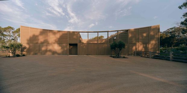 A low, minimalist contemporary building viewed from a broad paved forecourt. The facade is composed of warm brown timber-toned cladding and dark framed glazing, arranged in long horizontal volumes around a recessed central entry or courtyard opening. The building sits within a leafy landscape with timber fencing and mature trees, under a bright sky.