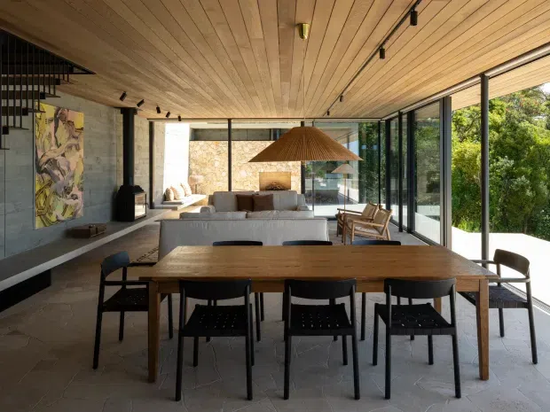 Open-plan contemporary dining and living space with a long timber dining table and black-framed dining chairs in the foreground. A large upholstered sofa, sculptural wood pendant light, fireplace, and lounge seating occupy the background. Full-height glazed walls open to dense greenery, while the room features a warm timber slatted ceiling, exposed concrete walls, stone flooring, and dark metal framing.