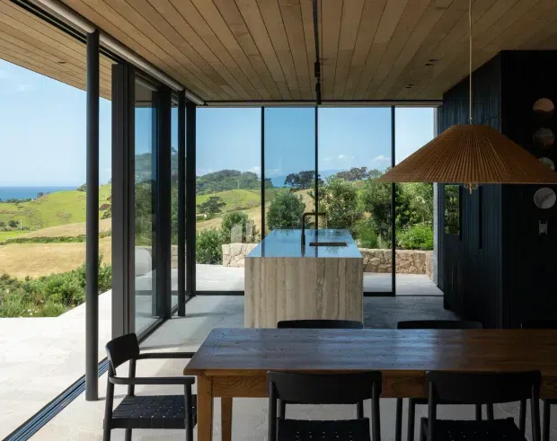 A refined open-plan dining and kitchen interior with a long timber dining table and black chairs in the foreground, a stone island bench beyond, and full-height black-framed glazing opening to a sunny coastal landscape. The room features a timber-lined ceiling, dark vertical wall joinery, polished concrete flooring, and a large woven pendant light.
