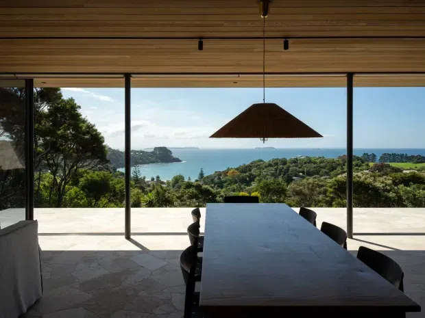 A contemporary dining area opens onto a broad terrace through full-height glazing, with a long dark dining table and black chairs beneath a large pendant light. The timber-lined ceiling, slender dark window frames, stone-paved floor and expansive coastal outlook create a refined indoor-outdoor setting.