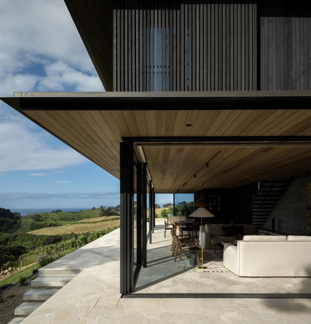 A contemporary multi-level coastal home viewed from an expansive outdoor terrace. Deep cantilevered roof planes frame a mostly open living area with full-height glazed sliding doors, warm timber soffit ceilings, dark vertical timber screening, a concrete stair, and neutral furnishings. The terrace overlooks rolling green landscape and the ocean beyond.