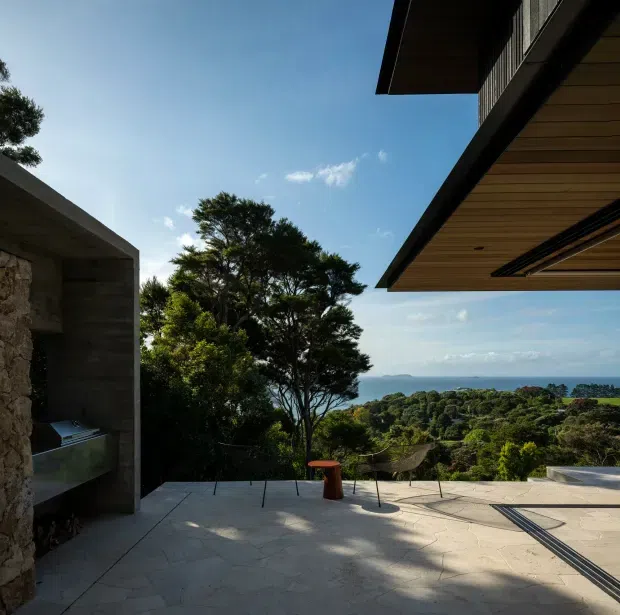 A contemporary outdoor terrace overlooking dense coastal vegetation and the ocean. The space features a large pale stone-paved terrace, a built-in concrete outdoor fireplace or barbecue structure at left, and broad cantilevered roof eaves with warm timber soffit lining at right. Minimal outdoor seating sits beneath mature trees, creating a refined indoor-outdoor setting with expansive coastal views.