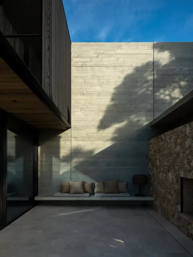 A narrow, sheltered architectural courtyard or outdoor lounge framed by dark timber-clad projecting volumes, a tall pale concrete wall, and a rough natural-stone wall with a built-in fireplace. A low built-in concrete bench with cushions provides seating beneath strong sunlight and tree shadows. The composition is minimalist and contemporary, with muted grey, charcoal, timber, and natural stone tones.