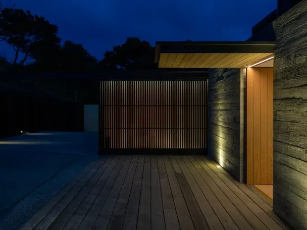 Twilight exterior view of a contemporary residential entry and garage area. A timber-lined deck leads to a tall vertical-slatted garage or privacy gate, beside a warm timber front door and dark textured masonry or concrete walls. Integrated lighting highlights the entry and wall surfaces, creating a dramatic, subdued atmosphere.