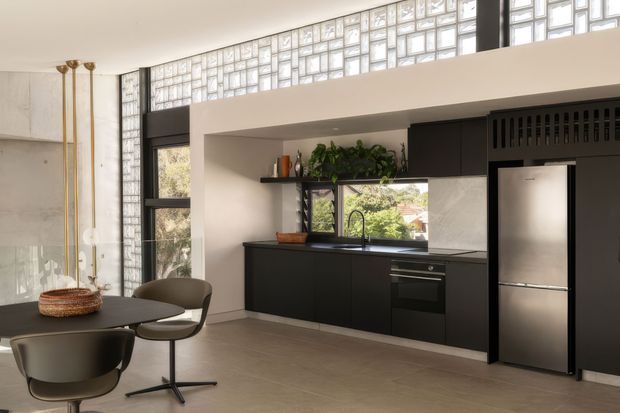 Contemporary open-plan kitchen and dining area with a sleek black built-in kitchen, stainless-steel refrigerator and oven, dark stone-look benchtops, and a small round dining table with sculptural chairs. A large steel-framed window and high band of translucent glass blocks bring in abundant natural light, while concrete and black-framed architectural elements add an industrial character.