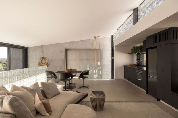 Open-plan modern interior combining a living room, dining area and compact kitchen. The space features a large beige sectional sofa, round dining table with dark sculptural chairs, polished concrete walls and floor, black built-in kitchen cabinetry, expansive dark-framed glazing, and a high clerestory band of textured glass blocks. A mezzanine-like upper edge and strong indoor-outdoor daylight contribute to the spacious, refined atmosphere.