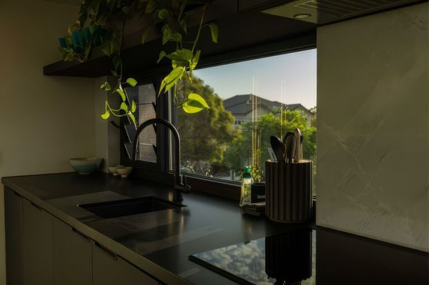 A moody contemporary kitchen vignette with dark cabinetry and a deep dark benchtop, a large black gooseneck tap over an undermount sink, and a reflective black cooktop or work surface. A large window brings in warm natural light and overlooks greenery and neighbouring homes. A trailing indoor plant hangs from an overhead shelf, while a ribbed cylindrical utensil holder and pale veined splashback add texture.