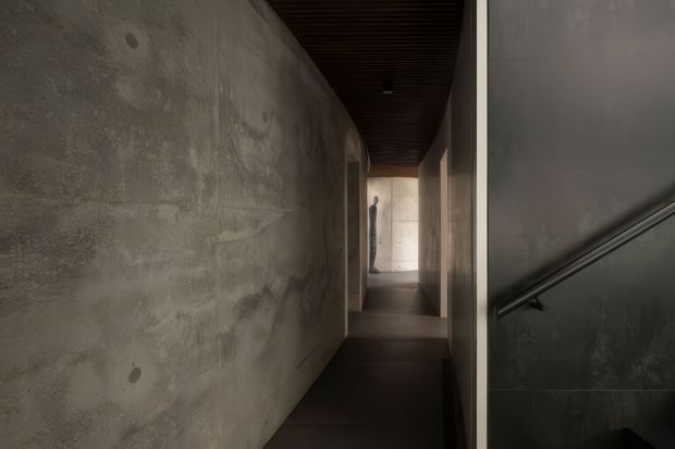 A narrow, dimly lit interior passage with a raw mottled concrete wall on the left, dark flooring, and a dark slatted ceiling. A stairwell with a simple metal handrail is visible on the right, while the corridor leads toward a brighter concrete-lined opening and a silhouetted figure in the distance. The restrained material palette and stark lighting create a moody, minimalist architectural atmosphere.