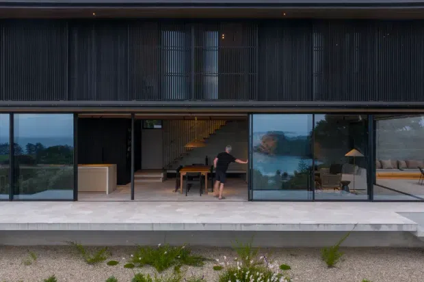 Exterior view of a dark, contemporary two-storey residence with extensive floor-to-ceiling glazing on the lower level and a screened upper facade. The broad terrace and landscaped gravel foreground frame views into an open-plan interior, where a dining area, staircase, kitchen joinery, and living space are visible. Strong horizontal lines, slim dark frames, and vertical screening create a refined modern architectural composition.