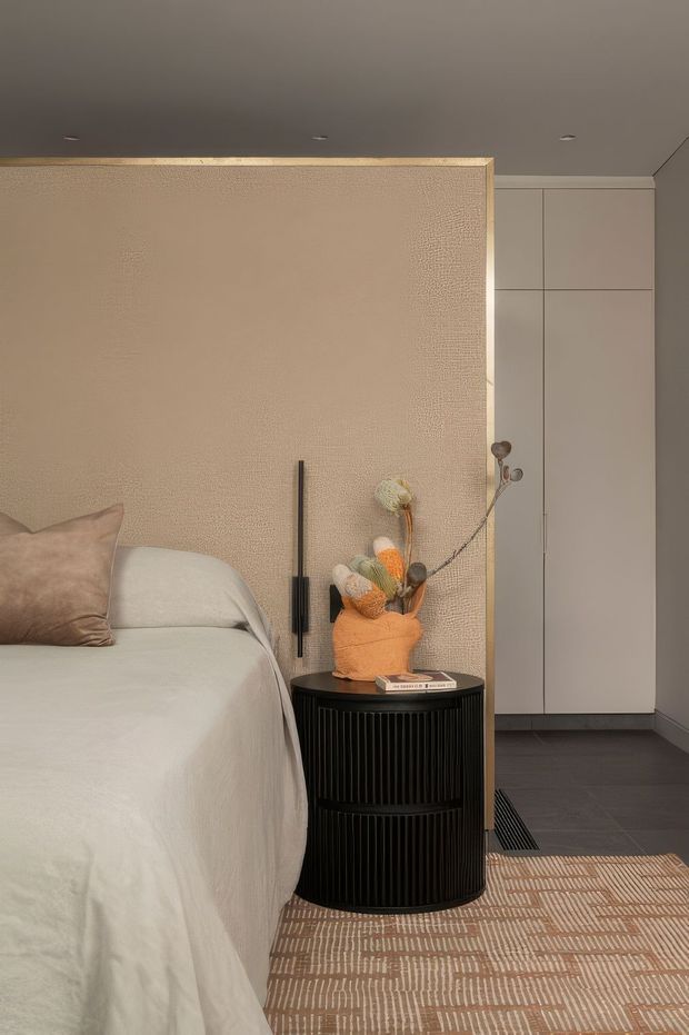 A warm, refined bedroom corner featuring a partially visible bed with white bedding and a taupe cushion, a textured beige upholstered or plaster-like feature wall with slim black wall lighting, a round black fluted bedside table, built-in pale cabinetry, dark timber flooring, and a patterned neutral area rug. Dried floral stems add a small decorative accent.