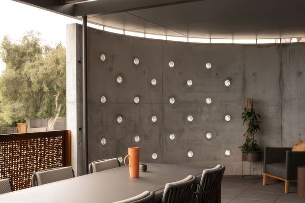 A contemporary dining and living area with a long light-coloured table, dark woven dining chairs, and a dark lounge chair. The space features a raw exposed concrete feature wall punctuated by regularly spaced circular inset lights, a broad curved clerestory window, a dark structural ceiling, and a perforated metal privacy screen opening toward leafy vegetation outside. A potted indoor tree and restrained furnishings create a refined industrial atmosphere.