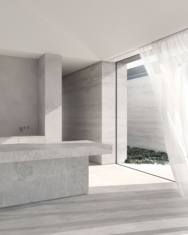 Minimalist, light-filled interior with a large monolithic grey stone island or counter in the foreground, smooth pale grey wall and stone surfaces, a full-height glazed opening, and sheer white curtains. A small planted courtyard is visible beyond the glazing. The space has crisp geometric architectural forms, abundant natural light, and a restrained neutral palette.