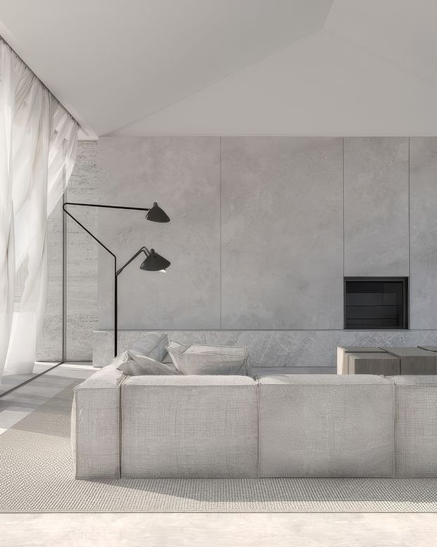 A bright, minimalist living room dominated by a large pale grey concrete or stone-look feature wall with integrated dark fireplace or media recess. A light neutral sectional sofa faces the wall, with a slim black floor lamp beside it. Sheer curtains and full-height glazing on the left bring in abundant daylight. The room has a restrained monochromatic palette, textured neutral flooring, and clean contemporary lines.