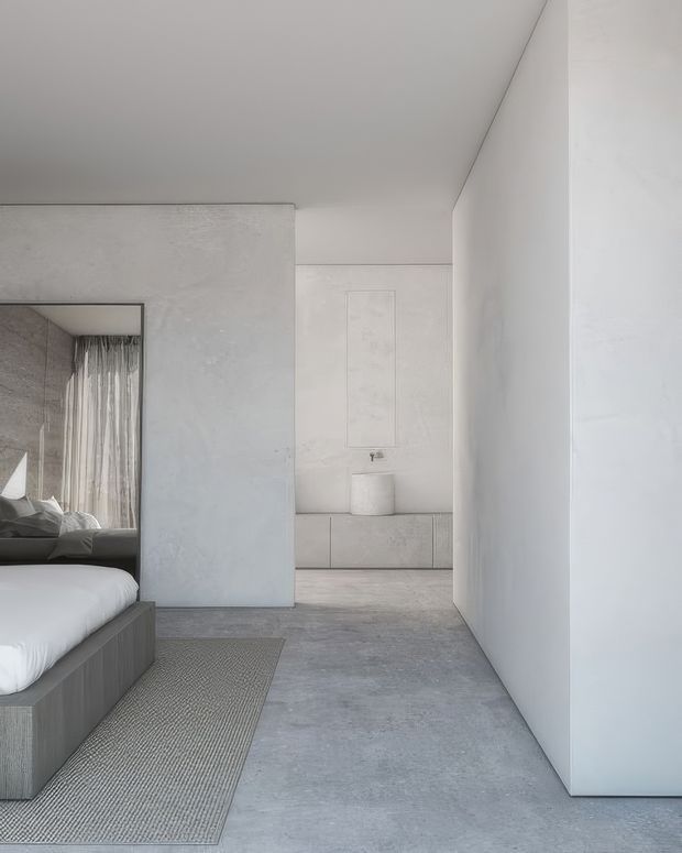 A minimalist bedroom interior viewed along a broad concrete-finished circulation space. The foreground shows a low dark built-in bed frame with white bedding and a textured neutral rug, while a large frameless opening reveals a secondary sleeping area or dressing zone with sheer curtains. Pale textured walls, flush cabinetry, a low built-in console, and a seamless grey floor create a restrained monochromatic scheme with strong clean lines and abundant daylight.