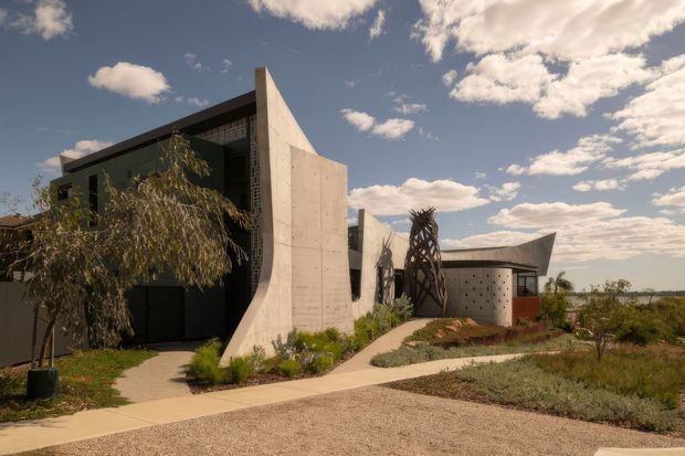Exterior view of a sculptural contemporary concrete house set within a landscaped, arid garden. The building features bold curved and angled concrete walls, dark recessed glazing, patterned breeze-block screening, and a low curved wing with a projecting roof form. Pale paths weave through native-style planting beneath a partly cloudy sky.