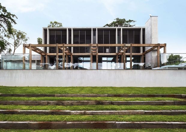 Exterior view of a modern multi-level building set behind a broad stepped lawn. The facade combines a rectilinear concrete volume, extensive dark louvred glazing, and a prominent timber post-and-beam pergola or screen across the lower level, with glass balustrades and an open terrace. The composition is clean and contemporary, with strong horizontal lines and integrated outdoor space.