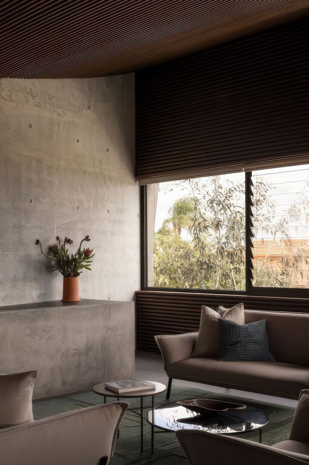 A moody, refined living room with a raw concrete wall and built-in concrete ledge, dark horizontal timber slatted ceiling and window surround, and expansive glazing overlooking trees. Neutral upholstered lounge chairs and a sofa are arranged around small round coffee tables, with restrained sculptural decor and a potted arrangement.