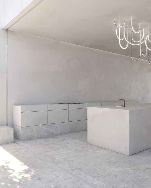 A serene, monochromatic minimalist kitchen with pale concrete or stone-look flooring, plastered walls, and seamless light-grey cabinetry. A large rectangular island with an integrated sink and dark tap dominates the foreground, while a sculptural white pendant light hangs overhead. The space is bright, quiet, and highly pared back, with subtle textured finishes and strong geometric forms.