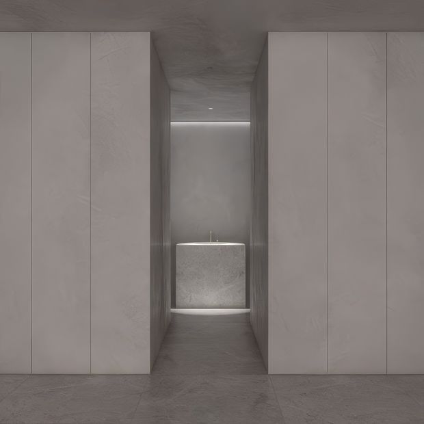 Minimalist monochromatic interior with full-height pale grey stone-look cabinetry or wall panels forming a narrow central passage. At the end is a compact rounded rectangular stone basin or vanity with a slim tap, softly illuminated by concealed lighting. Matching grey flooring and ceiling create a seamless, sculptural, gallery-like bathroom or dressing-area composition.