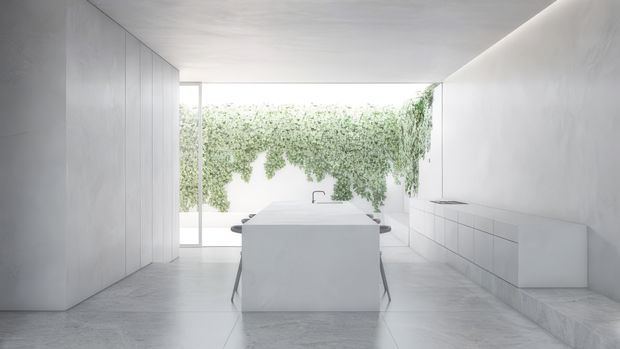 A highly minimalist white kitchen with a large central island, integrated handleless cabinetry along the right wall, full-height pale wall panelling on the left, and a broad opening to a lush vertical garden or courtyard. The space has a seamless light-toned floor, ceiling, and walls with concealed or minimal detailing and abundant diffused daylight.